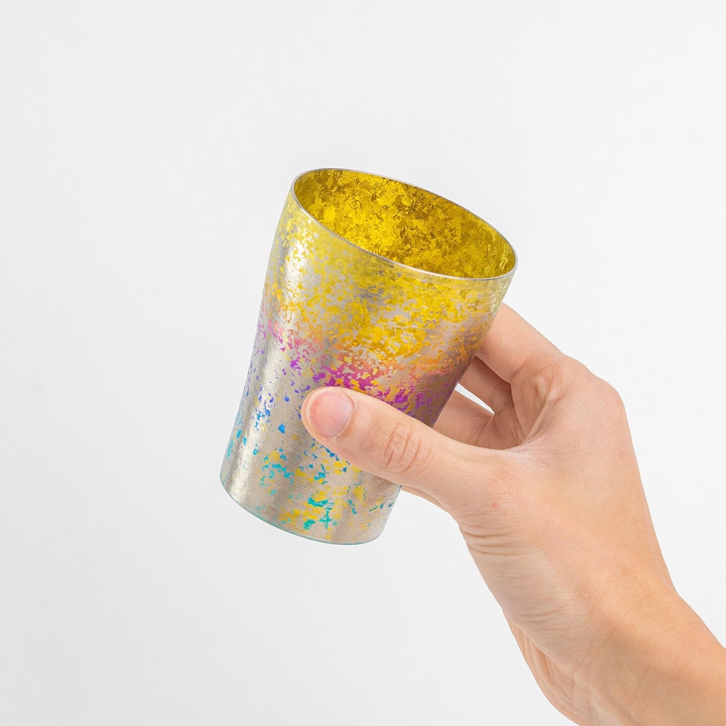 Hand holding the titanium tumbler at an angle, showing the golden yellow interior and rainbow exterior.
